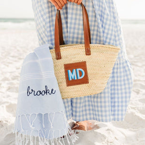 A girl in a blue and white gingham dress holds a straw tote with a blue monogram which holds a blue Turkish towel with a script navy name embroidered on it.