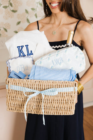 Woman holding a wicker gift basket filled with blue monogrammed essentials, striped towels, and a wine bottle.