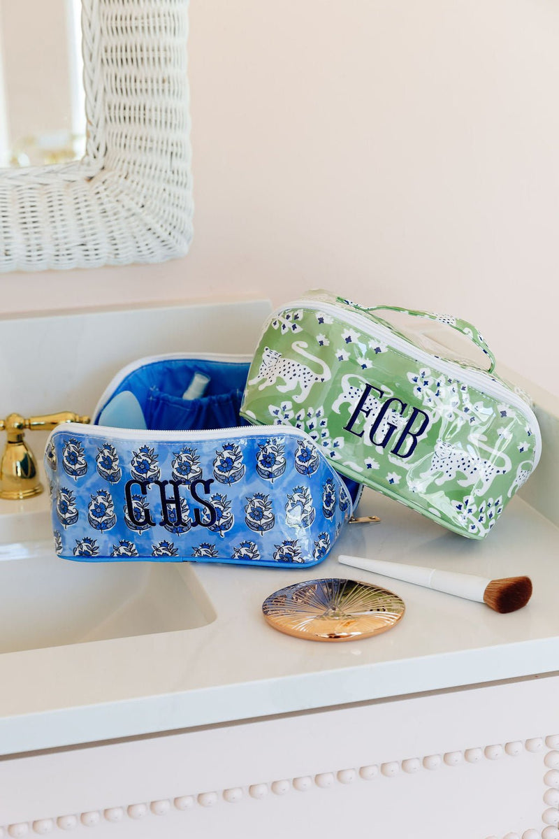 A blue and a green clear-coated flat lay pouch, monogrammed 'GHS' and 'EGB', on a bathroom counter with a makeup brush