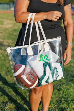 Close-up on grass: Gameday Clear Tote Bag stuffed with a football, green pom-poms, and a custom-initial water bottle.