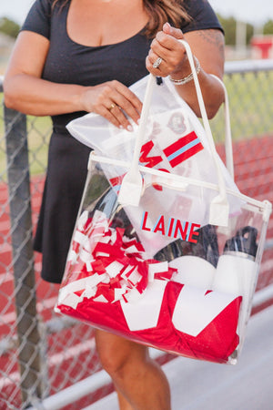 Woman at the track holds a Gameday Clear Tote Bag packed with red pom-poms and gear, personalized with angled “LAINE.”