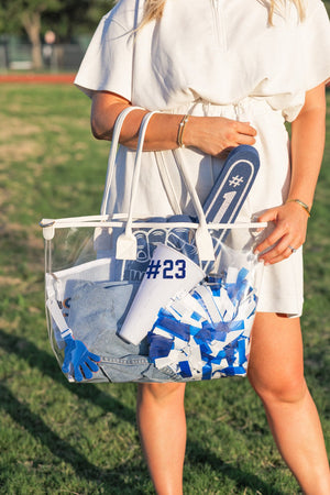 Gameday Clear Tote Bag carried by woman in white, filled with blue accessories and a mesh pouch printed “#23.”