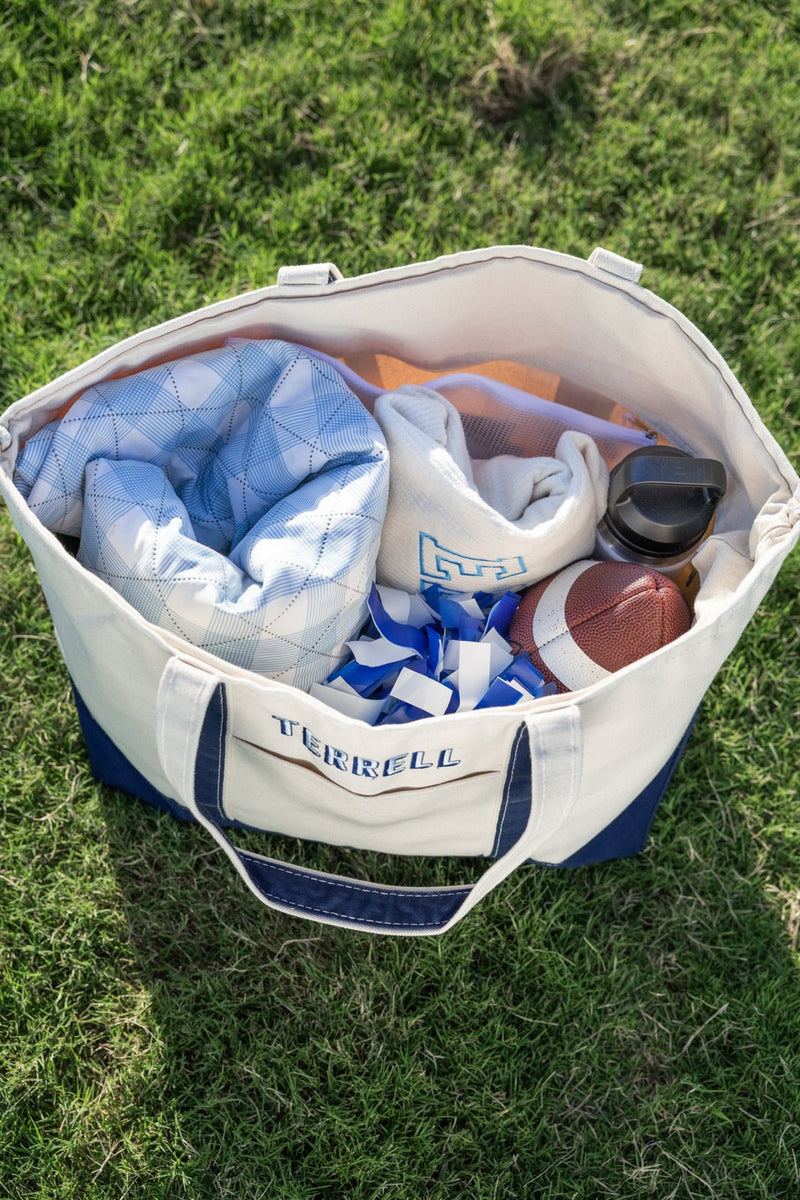 Open natural canvas tote with navy trim and embroidered “TERRELL,” filled with gameday gear (blanket, football).