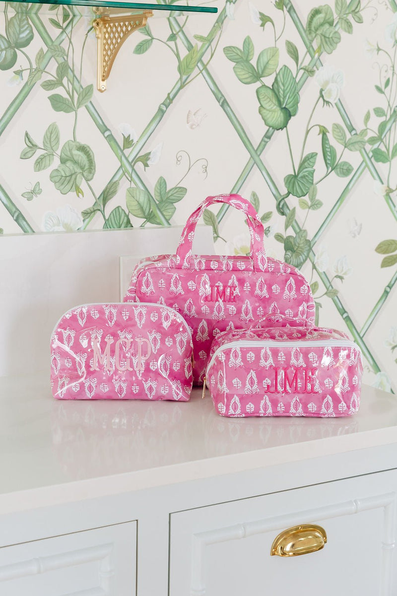 Three pink block-print bags: a hanging toiletry bag monogrammed 'JML', a dome pouch monogrammed 'MCP', and a rectangular pouch monogrammed 'JMB'