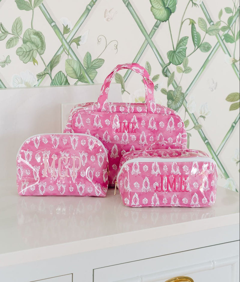 Three pink block-print bags: a hanging toiletry bag monogrammed 'JML', a dome pouch monogrammed 'MCP', and a rectangular pouch monogrammed 'JMB'