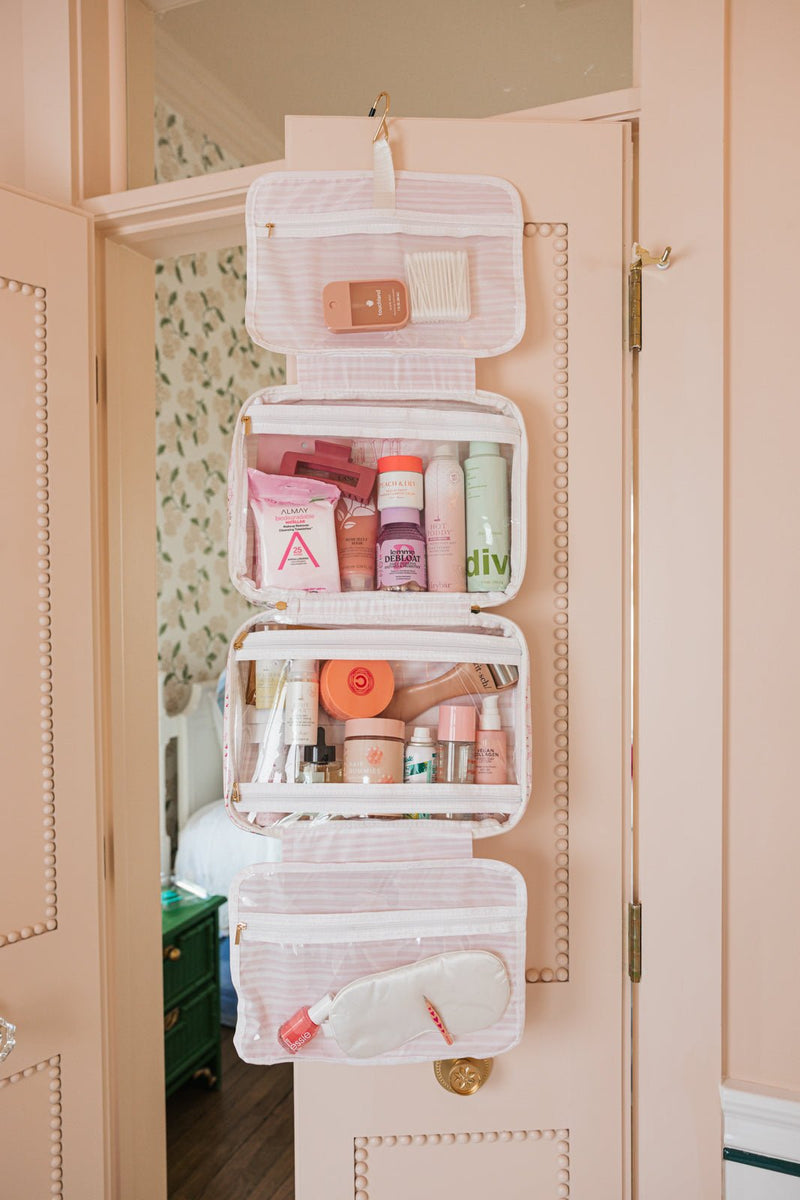 A hanging toiletry bag on a door with four clear compartments filled with skincare and makeup products