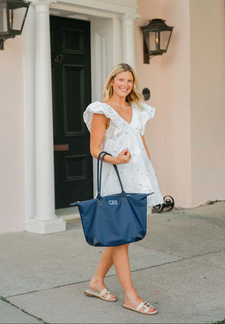 Woman in a white dress carrying a navy tote monogrammed "CHS" in white, walking outside a front door