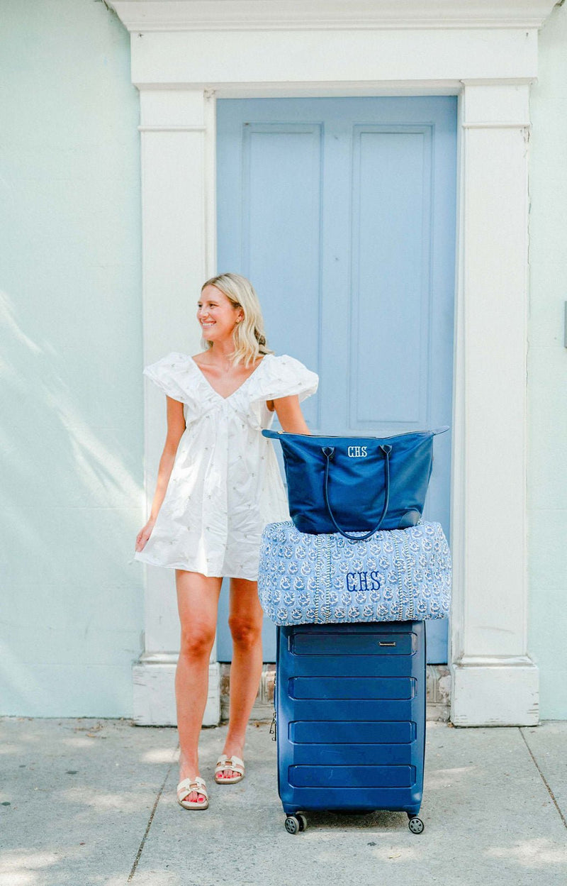 Woman standing with a navy tote monogrammed "CHS" and a blue block-print duffel monogrammed "CHS" stacked on a navy suitcase