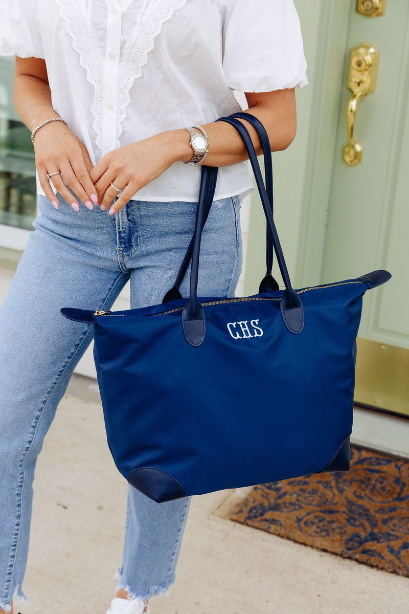 Close-up of a woman carrying a navy tote monogrammed "CHS" in white, walking outside a front door