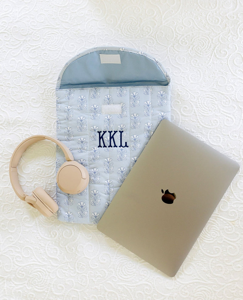 A personalized light blue quilted laptop sleeve with 'KKL' embroidered in navy, styled on a white textured blanket alongside a closed silver laptop, beige over-ear headphones, and a matching beige pouch.