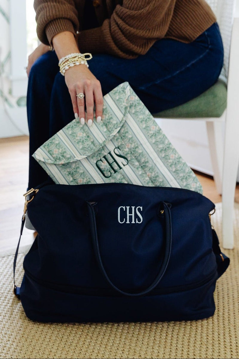 A navy blue personalized duffel bag with white monogrammed initials 'CHS' sits on a woven rug in a home setting, with a person sitting on a chair partially visible behind it placing a green patterned envelope or mailer into the bag.