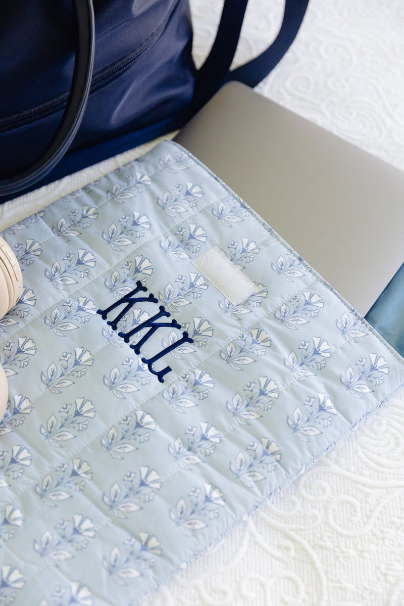 A light blue quilted monogrammed laptop sleeve with a blue floral pattern and the monogram 'KCL' embroidered in navy, styled on a white textured blanket next to a navy tote bag and a silver laptop.