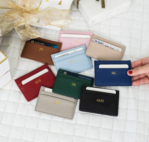 Nine leather cardholders in assorted colors with gold foil monogram initials, arranged on a quilted surface with gift boxes