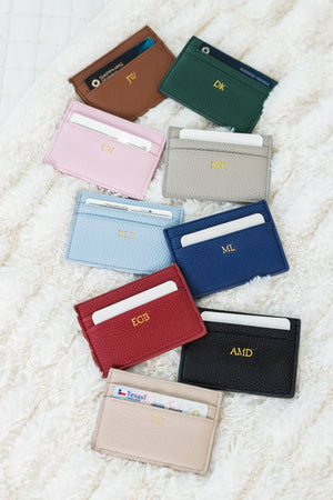 Nine leather cardholders in assorted colors with gold foil monogram initials, arranged on a fuzzy white rug