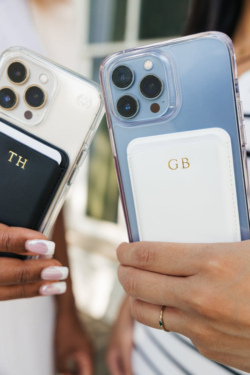 An assortment of smartphones with personalized card holders displayed on a pink and white checkered blanket. The collection includes various colors and monograms: black with 'AL', white with 'KD', navy with 'J', tan with 'MD', white with 'GB', black with 'TH', and navy with 'MF'. This vibrant display showcases the customizable options available for a stylish and practical phone accessory.
