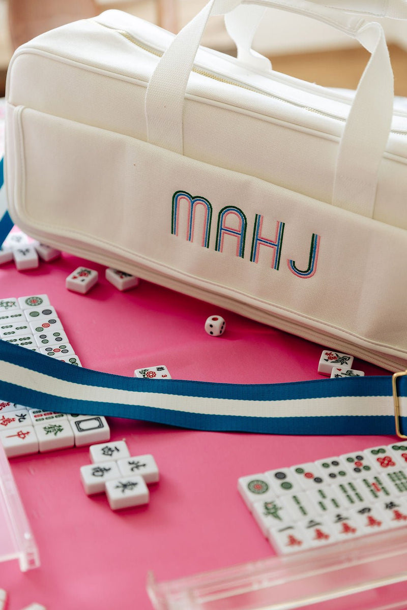 Close-up of "MAHJ" embroidery on a white canvas carryall duffel with a blue and white striped strap, surrounded by mahjong tiles and a die