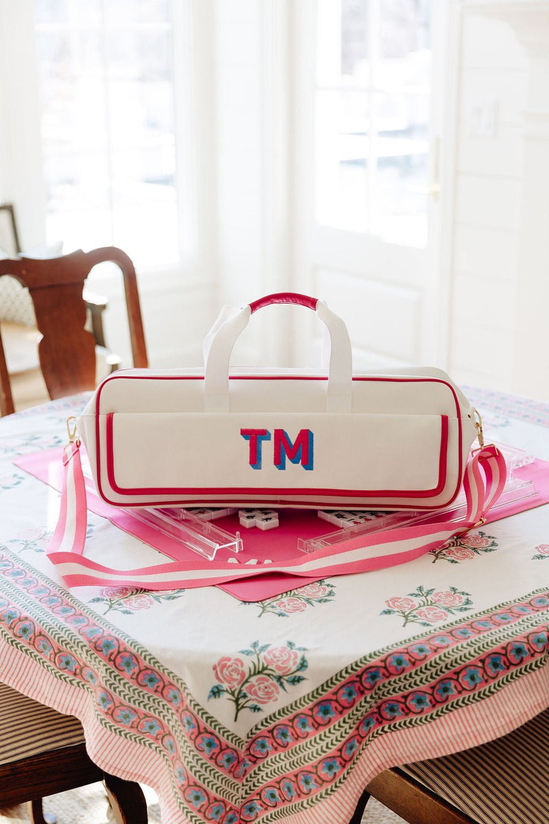 White canvas mahjong carryall duffel monogrammed "TM" sitting on a pink mahjong mat with tiles, on a floral tablecloth