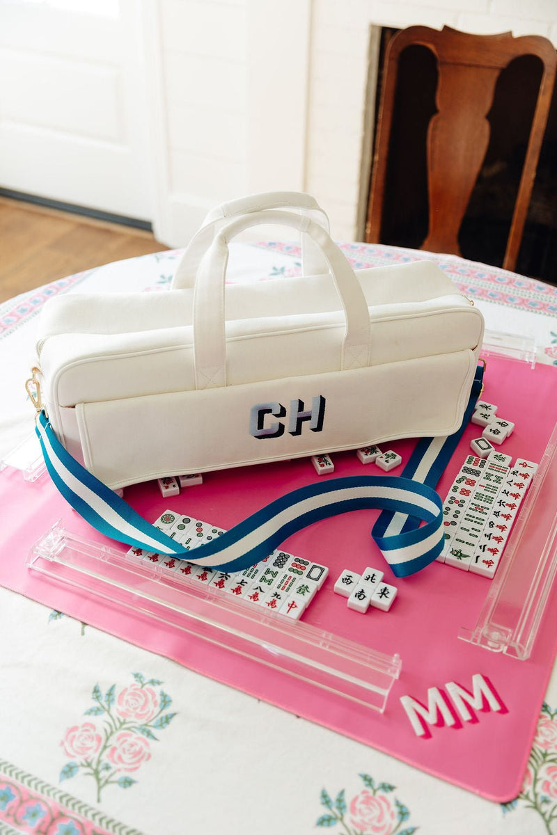 White canvas mahjong carryall duffel monogrammed "CH" with a blue and white striped strap, on a pink mahjong mat with tiles monogrammed "MM"