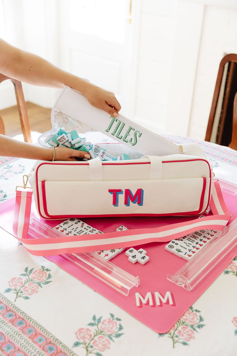 Hand unzipping a white mahjong carryall duffel embroidered "TILES" and monogrammed "TM," with mahjong tiles visible inside, on a pink mat monogrammed "MM"