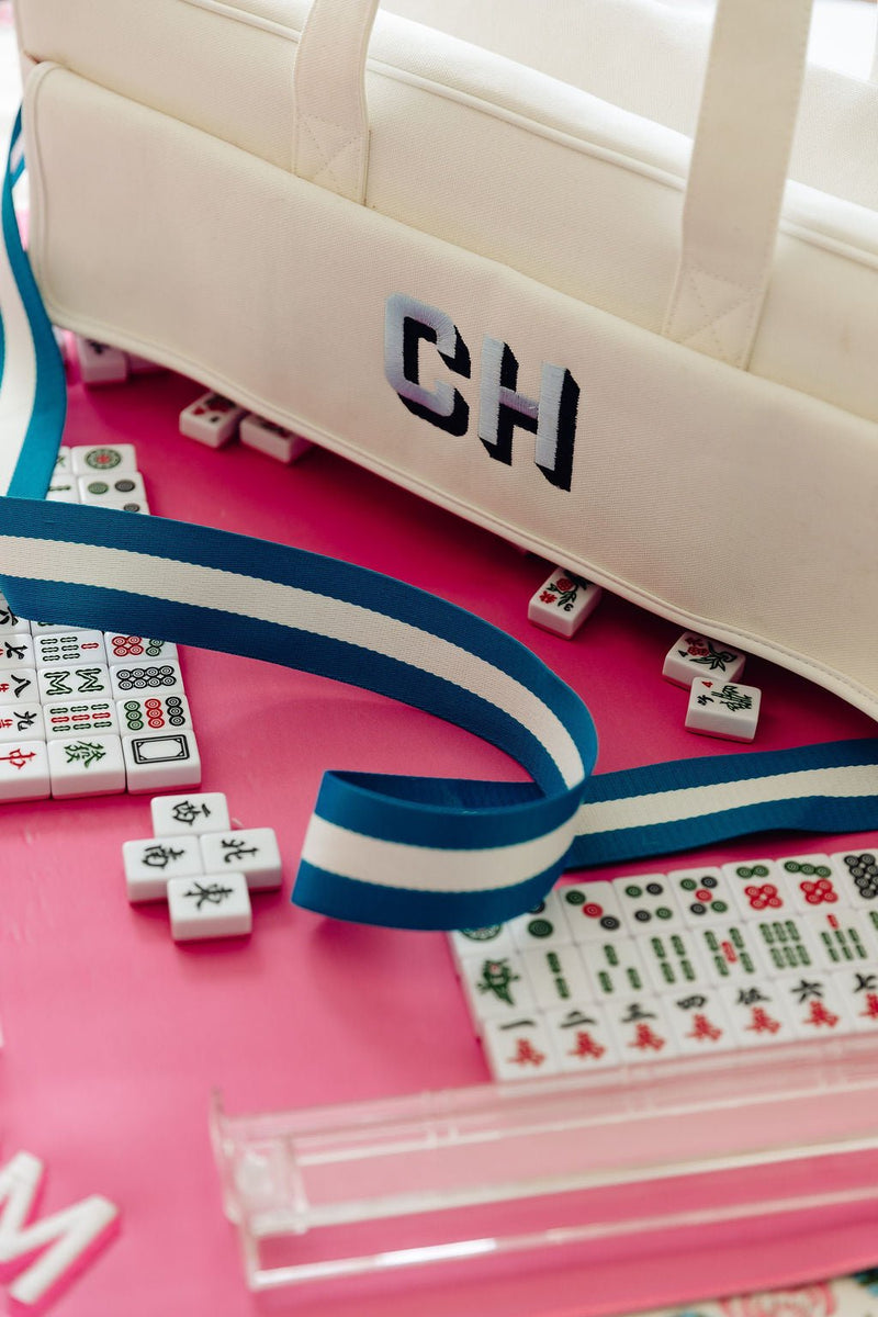 Close-up of "CH" monogram on a white canvas carryall duffel with a blue and white striped strap, surrounded by mahjong tiles