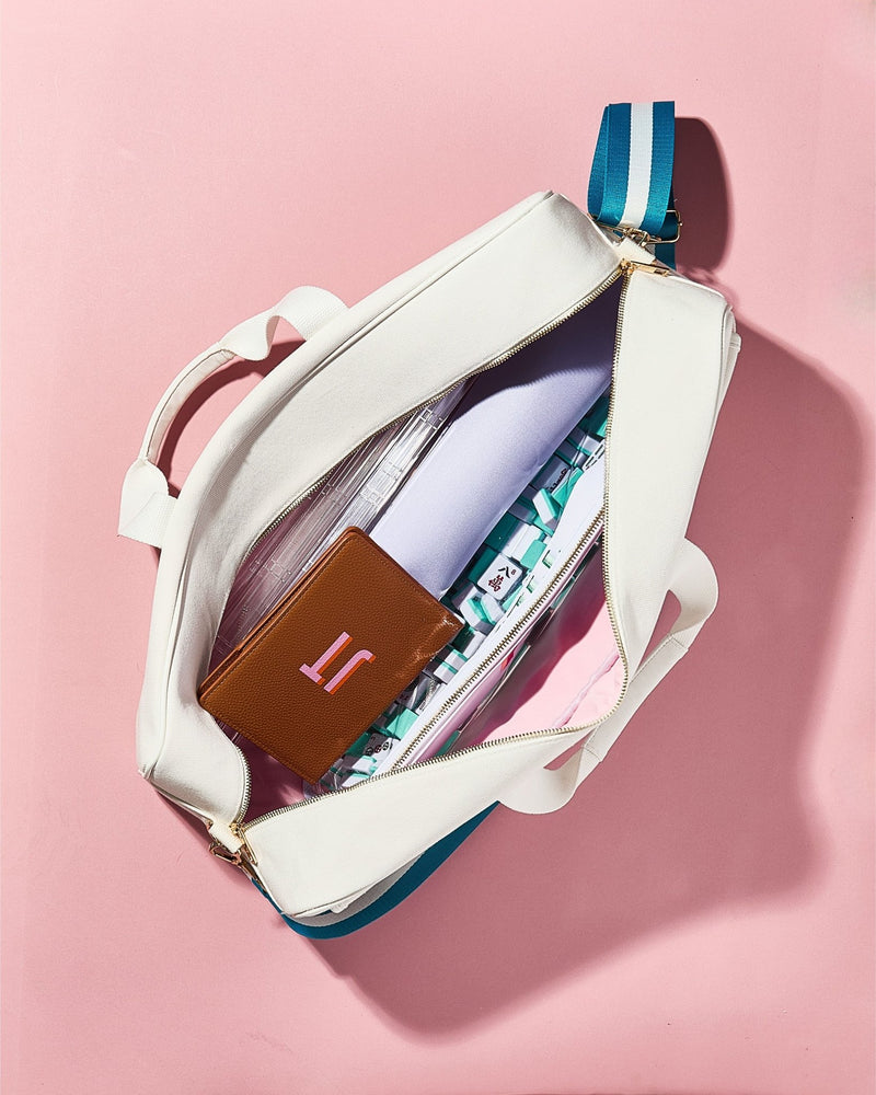 Overhead view of an open white canvas mahjong carryall duffel with a tan leather cardholder monogrammed "H" tucked inside, on a pink background