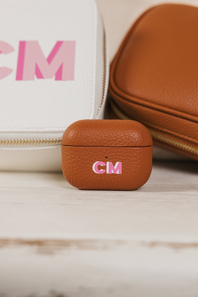 A personalized tan leather AirPods case with the initials "CM" in pink, showcased alongside a matching travel accessory set. The set includes a white monogrammed travel jewelry case and a tan leather tech organizer, creating a cohesive and stylish collection for on-the-go organization. The elegant personalization and high-quality materials make this set a perfect gift or travel essential.