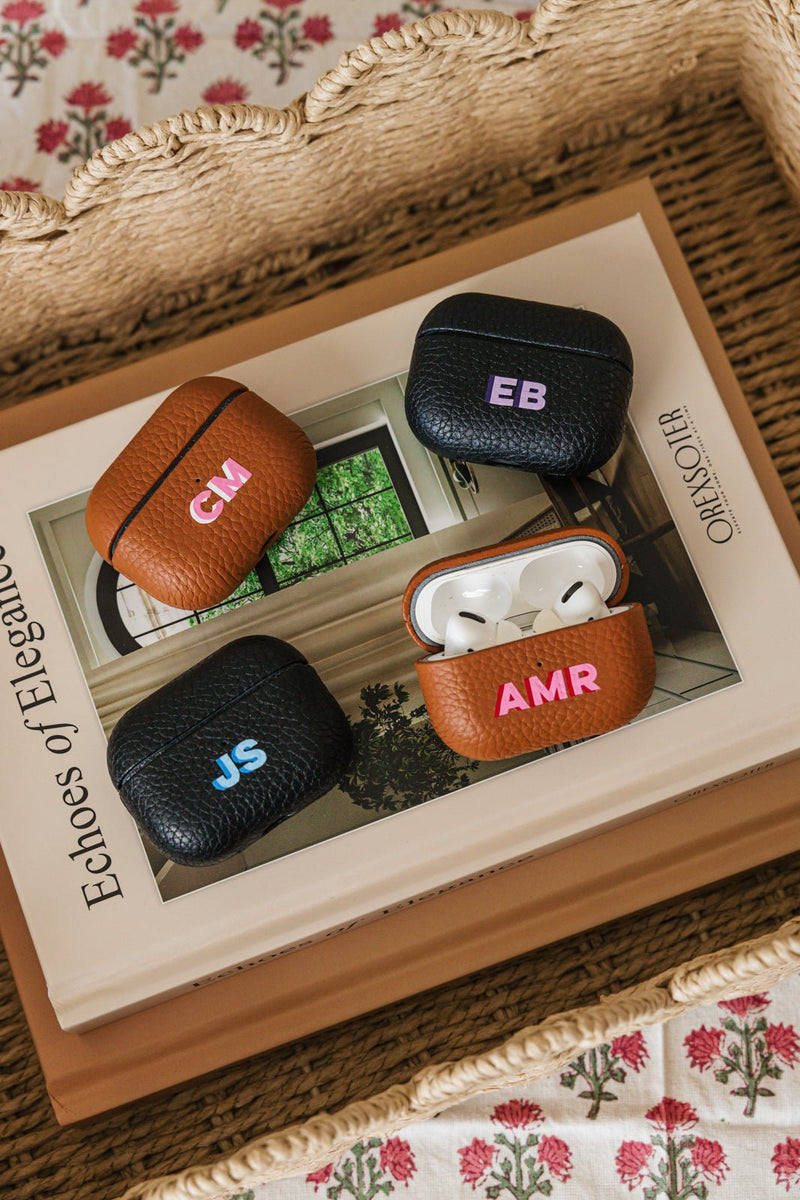 A beautifully arranged set of personalized leather AirPods cases displayed atop an elegant coffee table book. The collection includes both black and tan leather cases, each monogrammed with initials in vibrant colors, such as "CM," "EB," "JS," and "AMR." One of the cases is open, revealing sleek white AirPods inside. The scene is set within a woven basket, adding a touch of warmth and texture, while a floral fabric beneath enhances the aesthetic. 