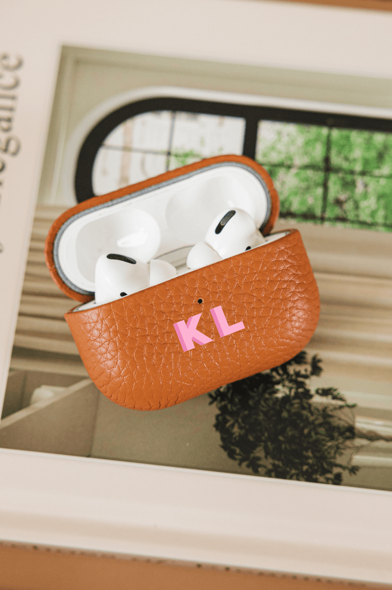 A stylish tan leather AirPods case, monogrammed with the initials "KL" in pink, rests on an elegant coffee table book, complementing a chic and personalized travel set. The case, part of a coordinated collection of monogrammed accessories, adds a sophisticated and functional touch, perfect for keeping tech essentials organized in style.