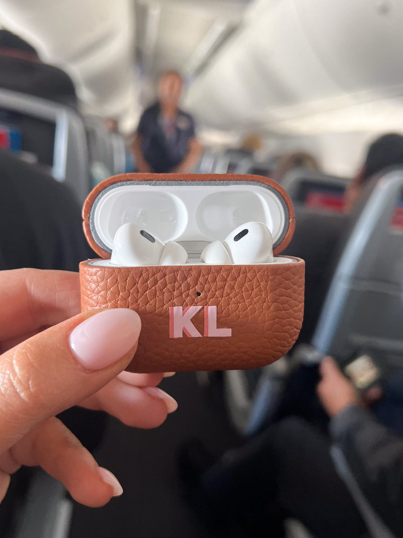 A stylish tan leather AirPods case, personalized with the initials "KL" in pink, is held mid-flight against the backdrop of an airplane cabin. This travel-ready accessory is part of a coordinated monogrammed set, perfect for keeping tech essentials organized while on the go. The luxurious leather texture and custom personalization make it both a practical and chic travel companion.