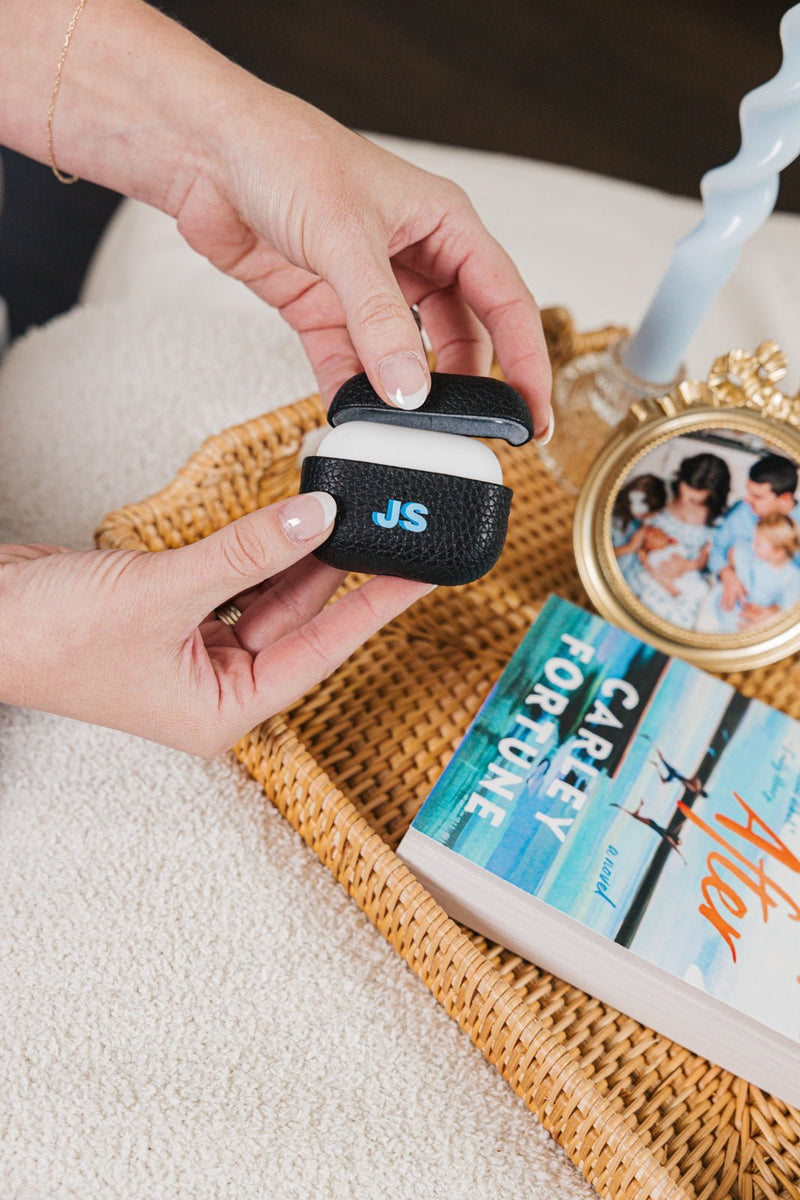 A sleek black leather AirPods case, personalized with the initials "JS" in blue, is gently held by a woman relaxing with a cozy setup. The case, part of a matching monogrammed travel set, rests on a woven tray alongside a book, a decorative candle, and a gold-framed family photo. This personalized accessory adds a stylish and practical touch, perfect for staying organized at home or on the go.