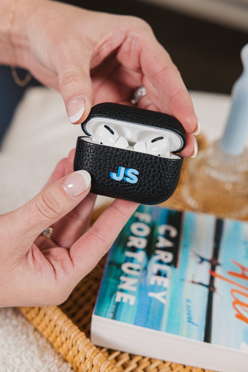 A stylish personalized black leather AirPods case with the initials "JS" in blue, part of a matching set of customized accessories. The case is held in a woman's hands, showcasing the sleek and protective design. A cozy reading setup featuring a book and woven tray in the background adds to the aesthetic, highlighting the perfect blend of functionality and personalization in this curated set.