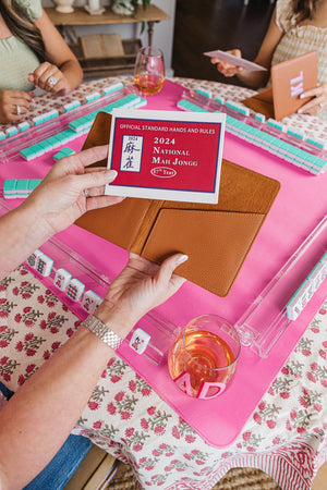 A stylish tan leather cardholder being used to hold a 2024 National Mah Jongg League rule card during a vibrant game night. The scene features a pink Mah Jongg mat, personalized acrylic racks, and monogrammed wine glasses, creating a coordinated and customized setup. The matching set of leather accessories and personalized drinkware adds a chic and sophisticated touch, perfect for a fun and stylish Mah Jongg gathering.