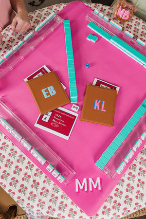 A vibrant and stylish Mah Jongg setup featuring a pink playing mat with personalized elements. Two tan leather cardholders, monogrammed with 'EB' and 'KL,' hold the official 2024 National Mah Jongg League rule cards, adding a chic touch to the game. The setup includes pastel blue tiles, clear tile racks, and a monogrammed wine glass labeled 'MB,' creating a coordinated and elegant gaming experience.