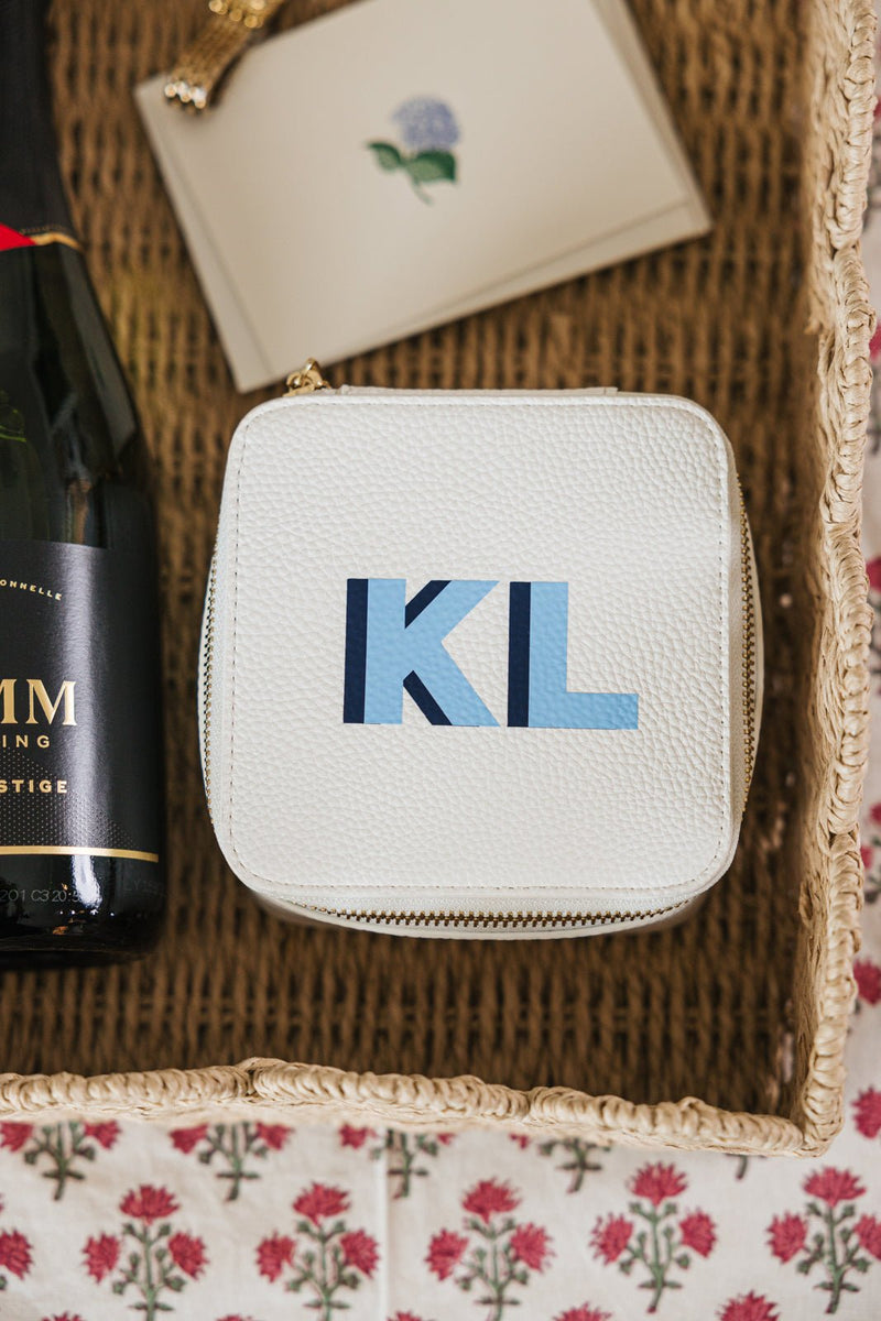 A personalized white jewelry case with the initials "KL" in a modern blue gradient design, placed inside a woven gift basket. The case features a zippered closure, perfect for organizing and protecting jewelry while traveling. Alongside it in the basket are a bottle of sparkling wine and a beautifully designed note card, making this a thoughtfully curated gift set ideal for special occasions. The floral tablecloth underneath adds a charming touch to the elegant presentation.