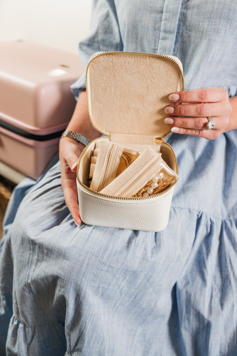A woman in a light blue dress elegantly holds an open personalized white travel jewelry case, showcasing its soft suede interior with organized compartments for rings, necklaces, and other accessories. The compact case, designed for travel, is filled with neatly stored jewelry in zippered pouches. A blush pink suitcase sits beside her, reinforcing the stylish and functional nature of this set as an essential travel companion. 