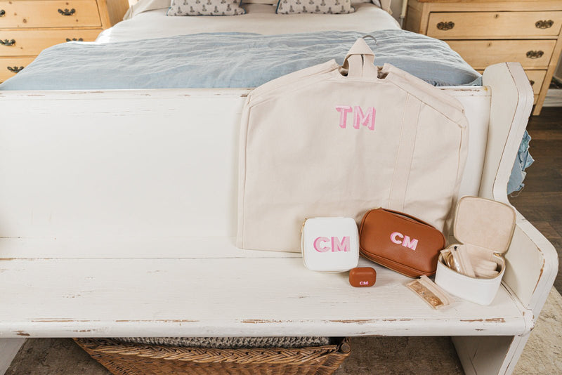 A beautifully curated personalized travel set displayed on a white wooden bench, featuring a monogrammed beige garment bag with "TM" embroidery, a brown leather travel case with "CM" initials, a white compact jewelry case with matching initials, and an open jewelry organizer with compartments for accessories. Also included is a coordinating monogrammed AirPods case and a delicate jewelry pouch.