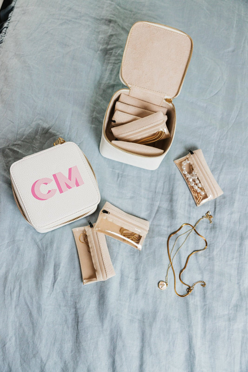 A beautifully organized personalized jewelry travel case set in white, featuring initials "CM" in pink on the exterior. One case is closed, highlighting its sleek design, while the other is open, revealing a soft beige interior with multiple compartments and zippered pouches for rings, necklaces, and earrings. Various gold and pearl jewelry pieces are displayed around the set, showcasing its functionality for keeping accessories tangle-free and secure. This stylish and compact set is perfect for travel and 