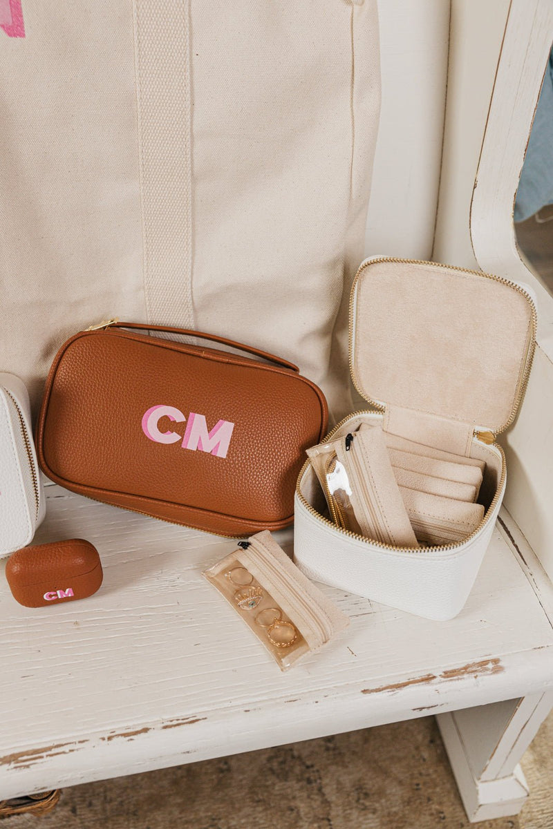  A beautifully curated personalized travel set displayed on a white wooden bench. The set includes a tan leather tech organizer with the initials "CM" in pink, a matching monogrammed white jewelry case with a zippered closure, and a coordinating small personalized AirPods case. The jewelry case is open, showcasing multiple soft velvet compartments designed for neatly storing rings, earrings, and necklaces. A beige garment bag with pink initials "CM" is propped against the bench, completing the set.