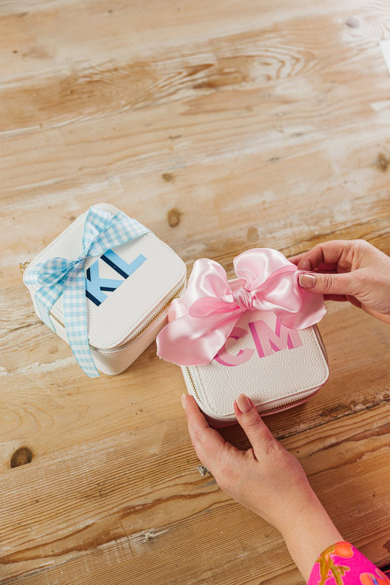 Alt text: A set of two personalized white travel jewelry cases with monogram initials "CM" in pink and "KL" in blue, elegantly wrapped with satin bows—one in pink and one in blue—on a rustic wooden table. The hands of a person wearing a vibrant pink outfit are delicately adjusting the pink-ribboned case, highlighting the thoughtful and stylish gift presentation. The cases feature a zippered closure, making them perfect for organizing jewelry while traveling.