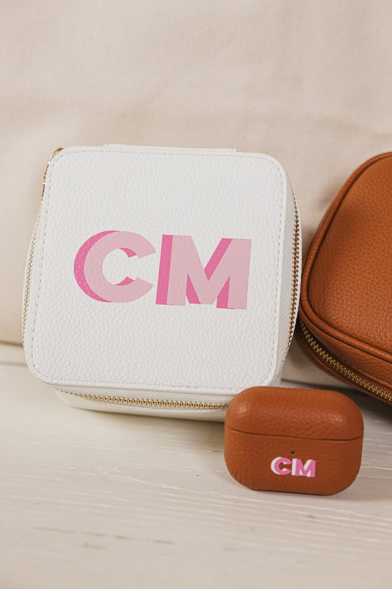 A stylish personalized travel accessory set featuring a white jewelry case, a brown tech organizer, and a matching monogrammed AirPods case, all customized with the initials "CM" in a pink gradient design. The set is displayed against a neutral background, showcasing the elegant and functional design ideal for travel and organization. The coordinated monogramming adds a chic, customized touch, making it a perfect gift or stylish storage solution for accessories.
