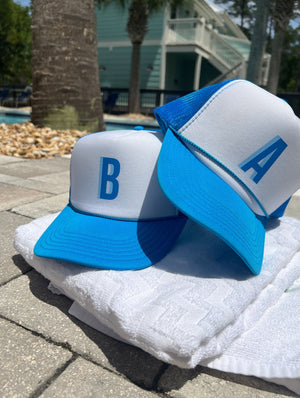 Two white trucker hats with blue brims and mesh backs, printed with blue letters 'B' and 'A', resting on a white waffle towel by a pool with palm trees in the background.