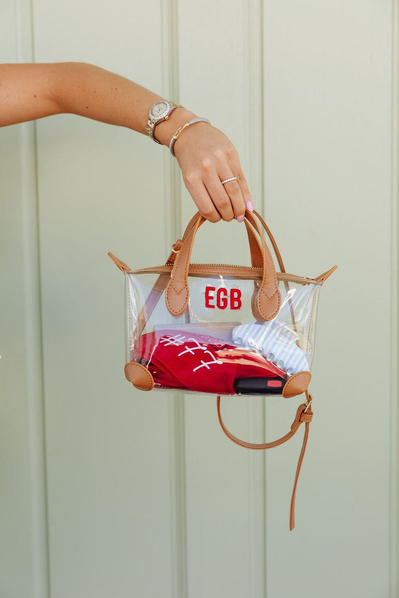Woman's arm holding a clear stadium mini crossbody bag with tan trim monogrammed "EGB" in red, containing a red pouch and striped fabric