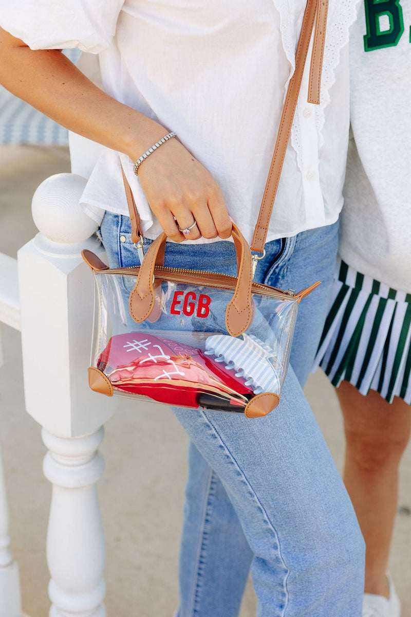 Close-up of a woman carrying a clear stadium mini crossbody bag with tan trim monogrammed "EGB" in red, standing next to someone in a green and white striped skirt