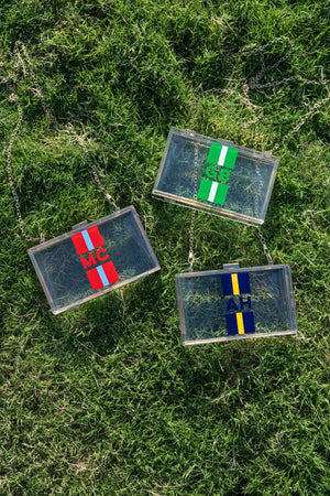 Three clear stadium bags on grass, each featuring vertical stripes and monograms—red “MC,” green “GB,” navy “AH”—with gold chain straps.