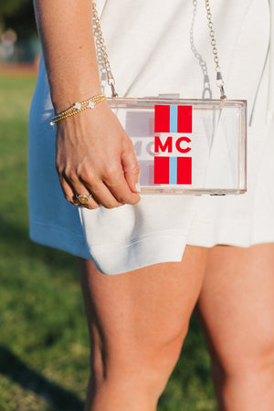 Clear stadium clutch with red and teal vertical stripes framing a bold “MC” monogram and gold chain strap.