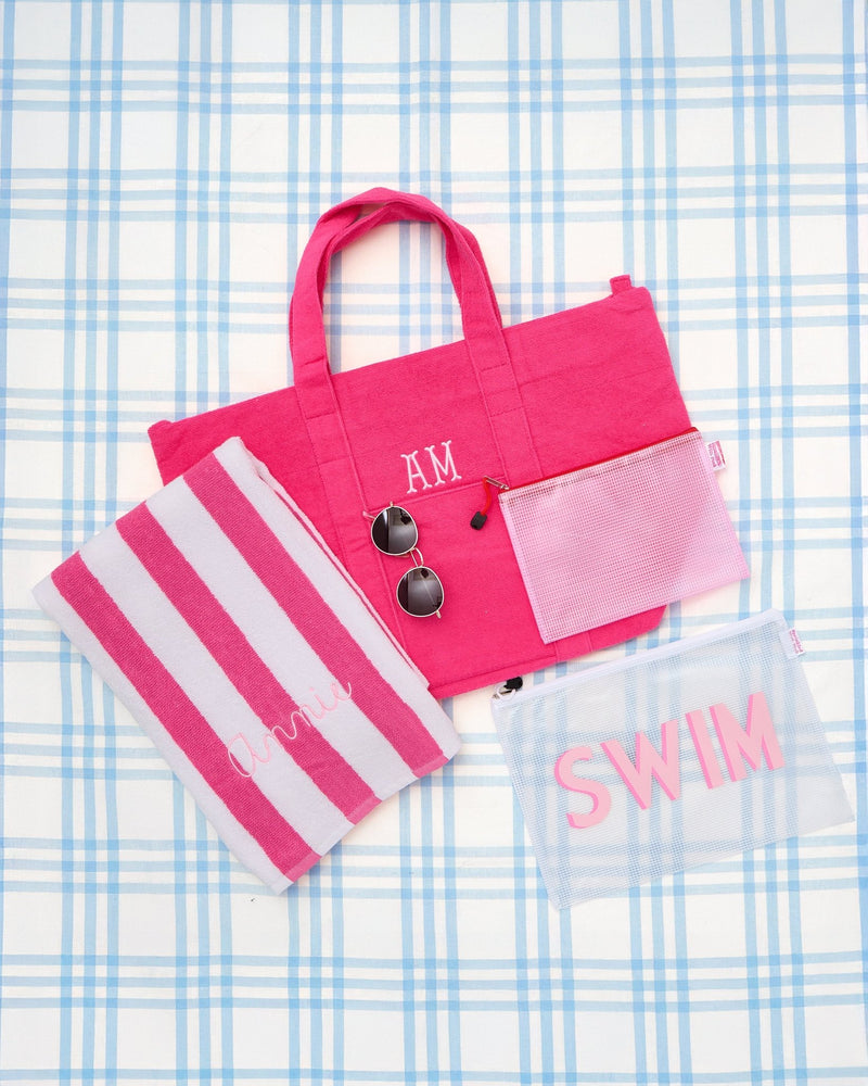 Pink swim gift bundle set featuring a personalized striped towel with 'Annie' embroidery, monogrammed terry cloth tote bag, pink mesh zip pouch, clear mesh pouch with 'SWIM' lettering, and sunglasses, styled on a blue plaid background—perfect for summer pool or beach gifting.