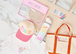 lat lay of teacher gift set including embroidered “TEACH” cap, “Mrs. Brown” sweatshirt, personalized water bottle, tote bags with embroidered names, and a clear pouch with “Mrs. Conder.”