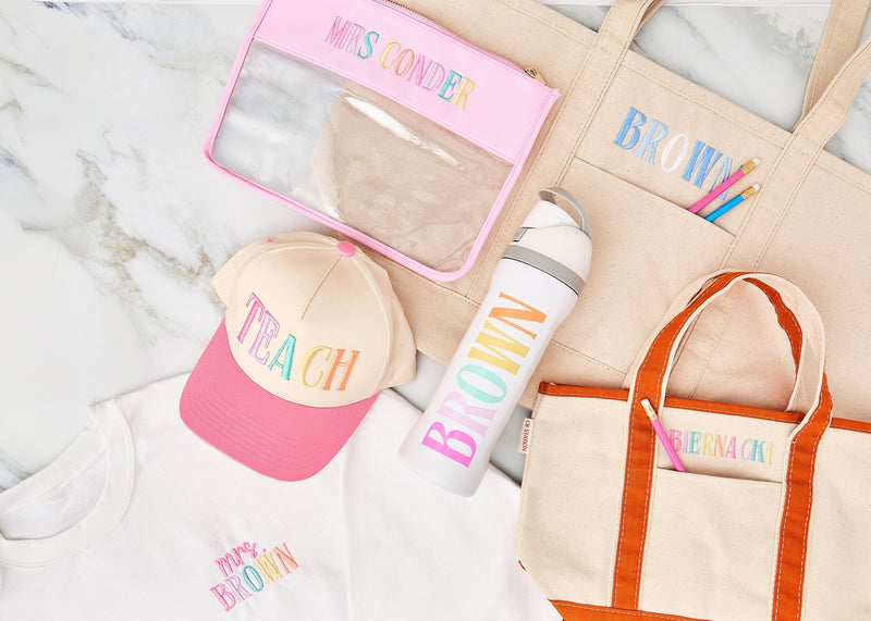 lat lay of teacher gift set including embroidered “TEACH” cap, “Mrs. Brown” sweatshirt, personalized water bottle, tote bags with embroidered names, and a clear pouch with “Mrs. Conder.”