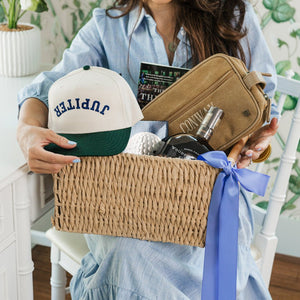 Woman smiling while holding a curated gift basket featuring a Custom Embroidered Upside Down Vintage Trucker Hat with “JUPITER” text, styled in a classic green and white color combo—perfect for a personalized and playful statement accessory.