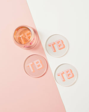 A wine glass sits on top of a coaster and next to three other matching coasters with a pink and melon block shadow monogram.